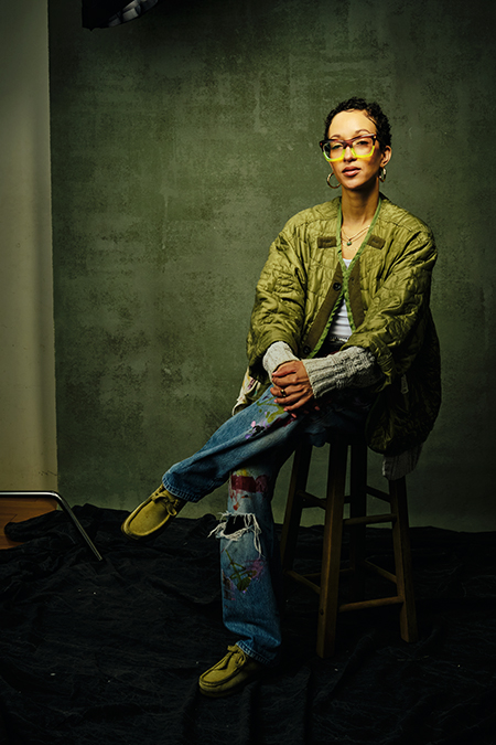 Artist Portrait from Being in New York Series Studio portrait of a female artist in a green jacket and paint-splattered jeans, seated on a stool in New York City.