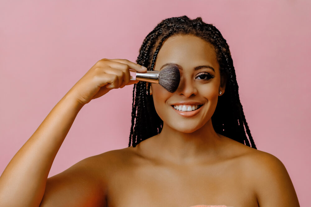 Young woman with dreadlocks smiling and covering one eye with a makeup brush during NYC personal branding studio portrait on a pink background